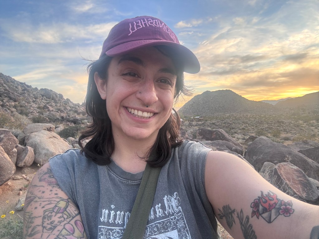 A selfie of Gia in front of a Rocky Mountain terrain and beautiful sunset in Joshua Tree. They are wearing a Blondshell baseball cap are are smiling